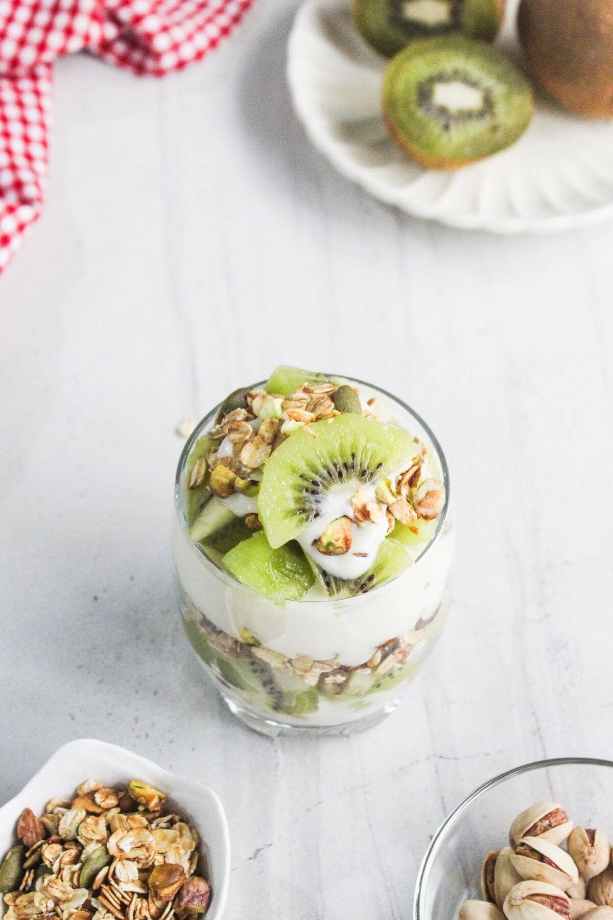 A glass filled with layers of yogurt, granola, and sliced kiwi. Sliced kiwi is also displayed on a plate in the background, next to a red checkered cloth.