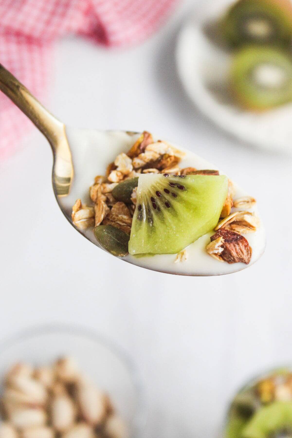 A spoonful of yogurt topped with granola, nuts, and a slice of kiwi fruit, held above a white surface with blurred background elements.