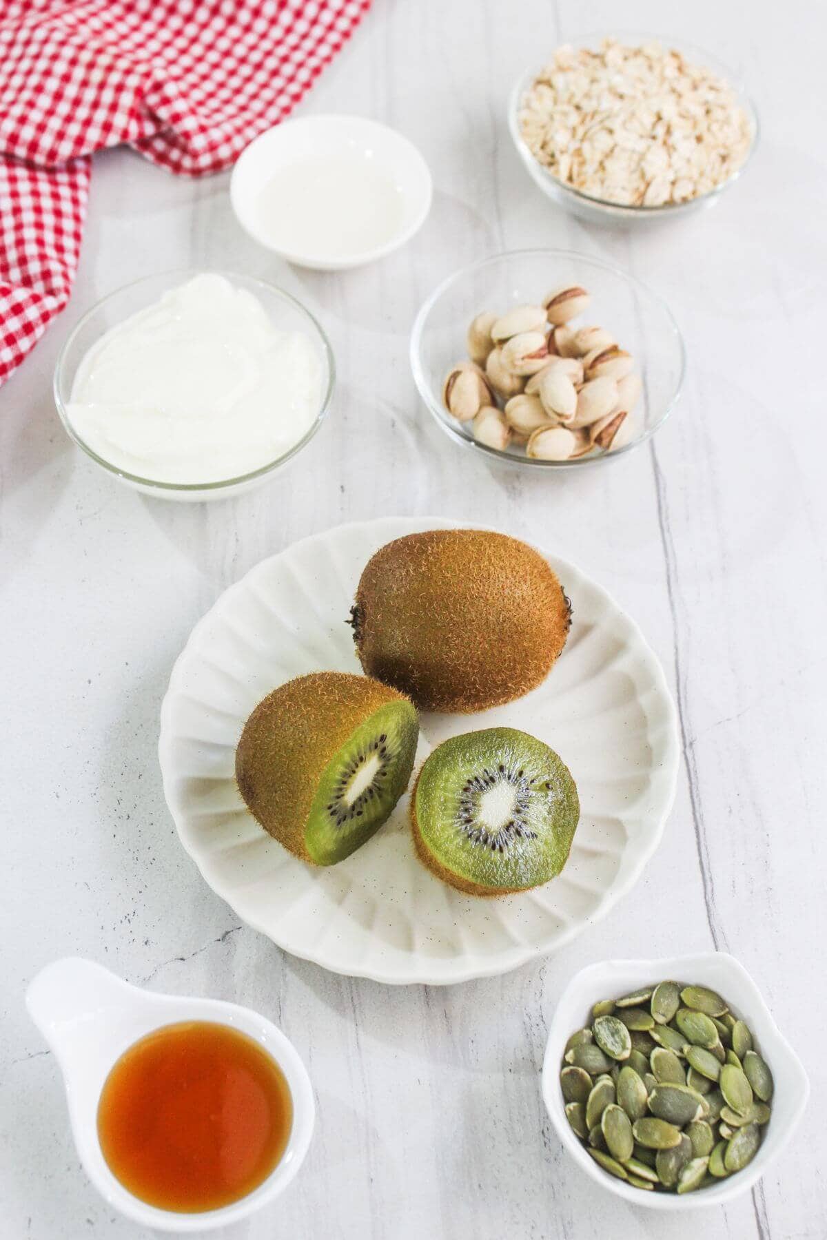 A plate with a whole and halved kiwi, surrounded by bowls of yogurt, oats, pistachios, pumpkin seeds, honey, and milk on a white surface.
