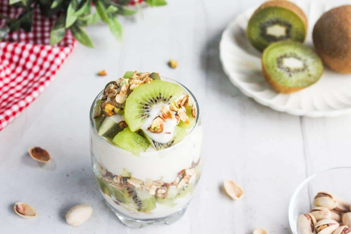A glass filled with layers of yogurt, granola, and kiwi slices is on a white surface. Pistachios and whole kiwis are nearby, along with a red checkered cloth and green leaves.