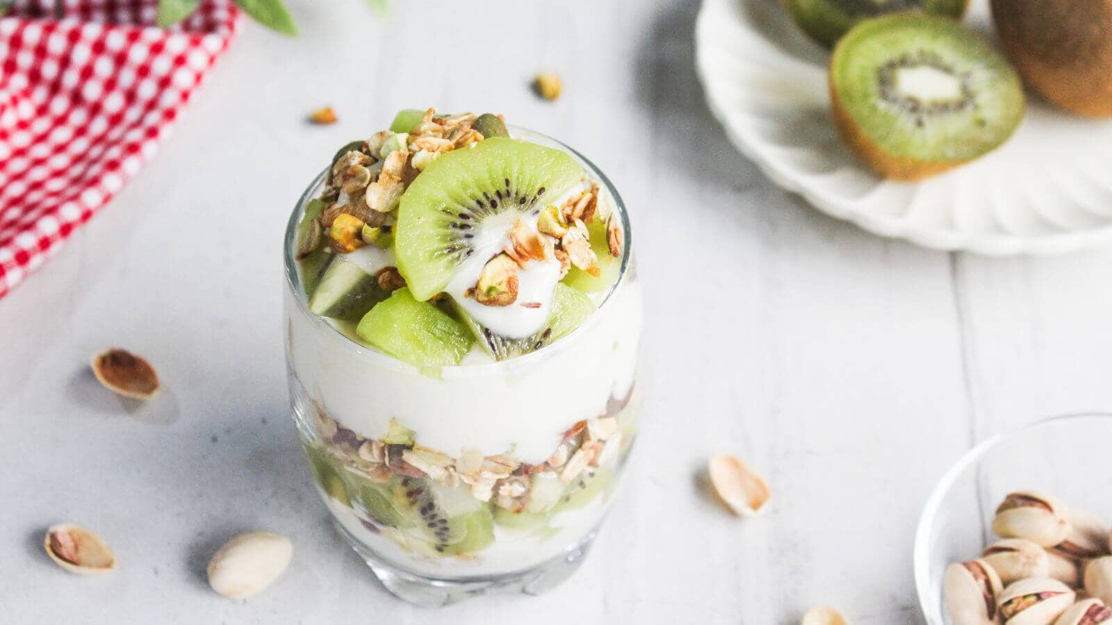 A glass parfait with layers of yogurt, sliced kiwi, and granola sits on a white surface, with pistachios and a plate of kiwis in the background.
