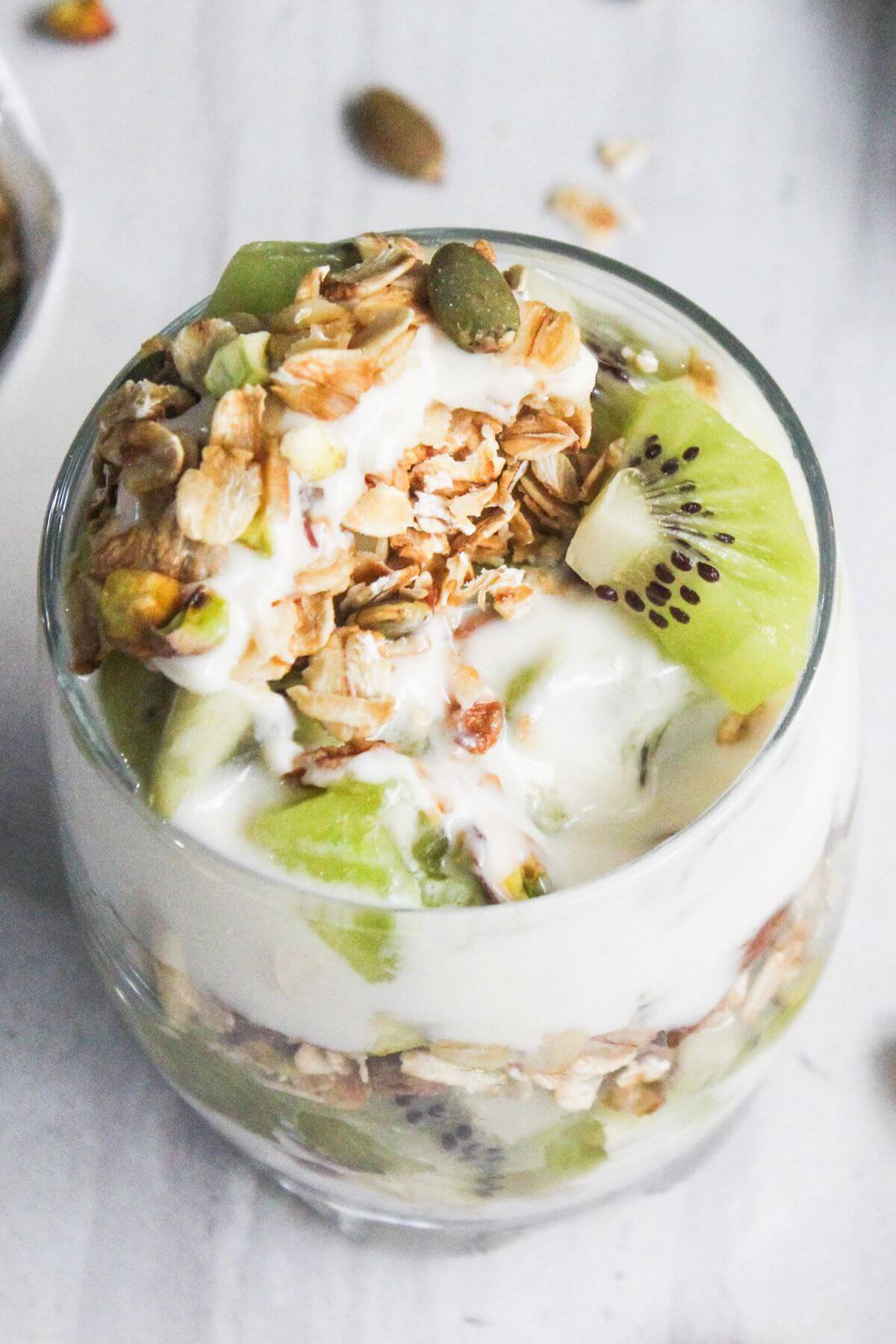 A glass filled with layers of yogurt, granola, and sliced kiwi, topped with granola and seeds, on a light-colored surface.