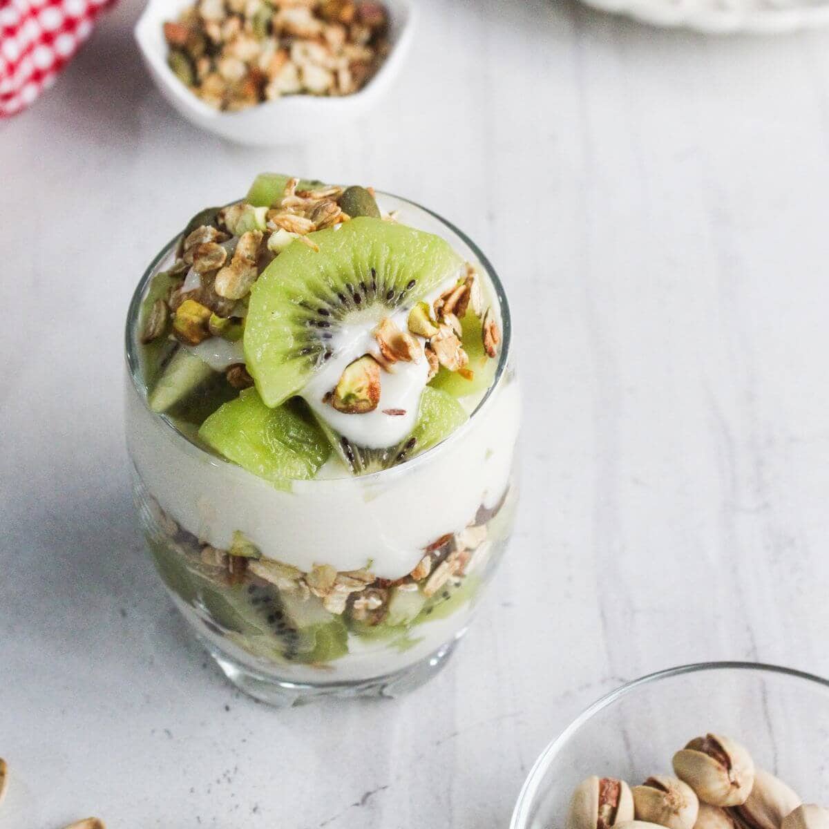 A glass filled with layers of granola, sliced kiwi, and yogurt sits on a white surface, with small bowls of granola and pistachios nearby.
