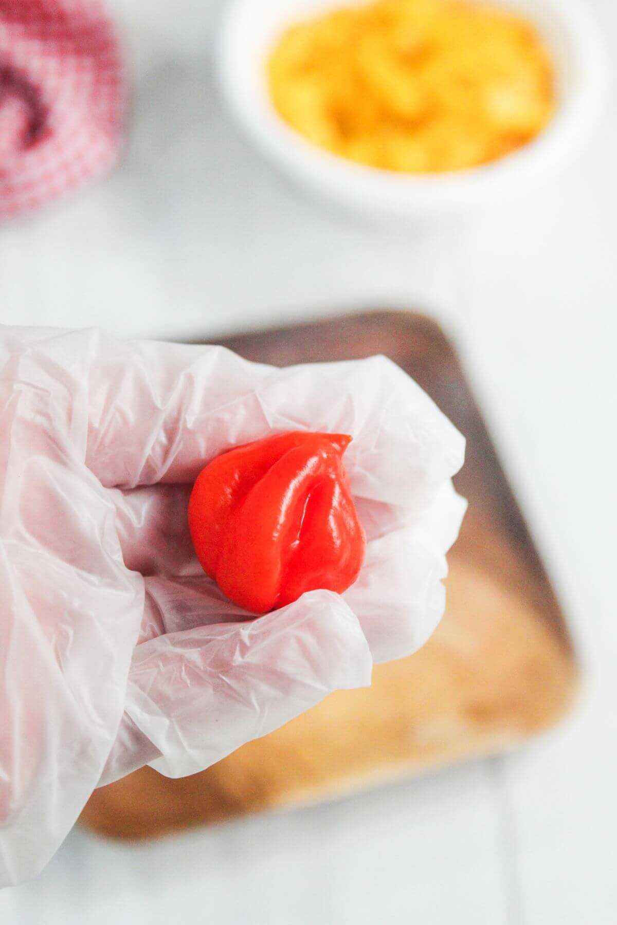 A gloved hand holds a bright red habanero pepper over a blurred background with a white bowl and wooden tray.