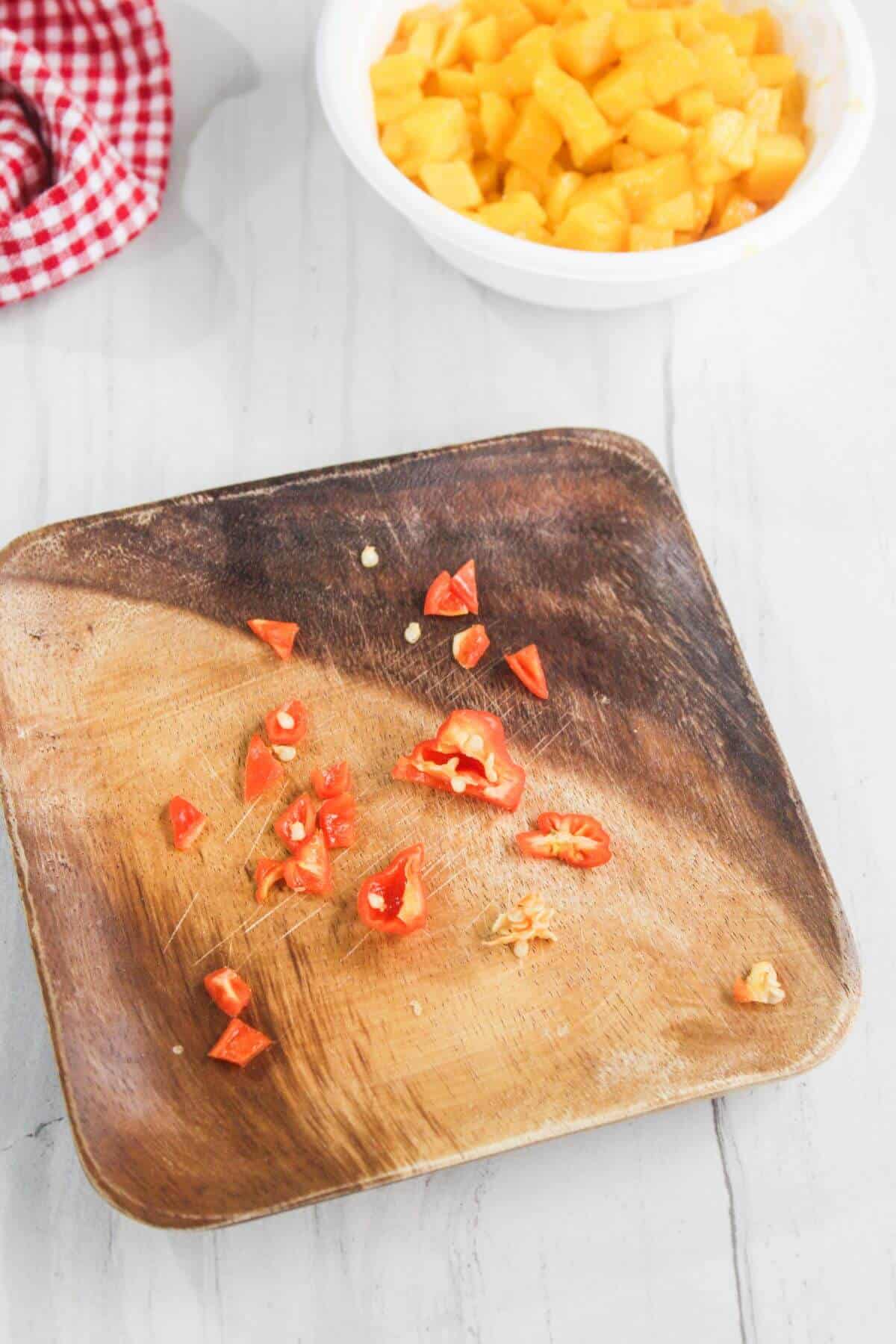 Diced red chili pieces on a wooden plate with a bowl of chopped mangoes in the background on a light countertop.
