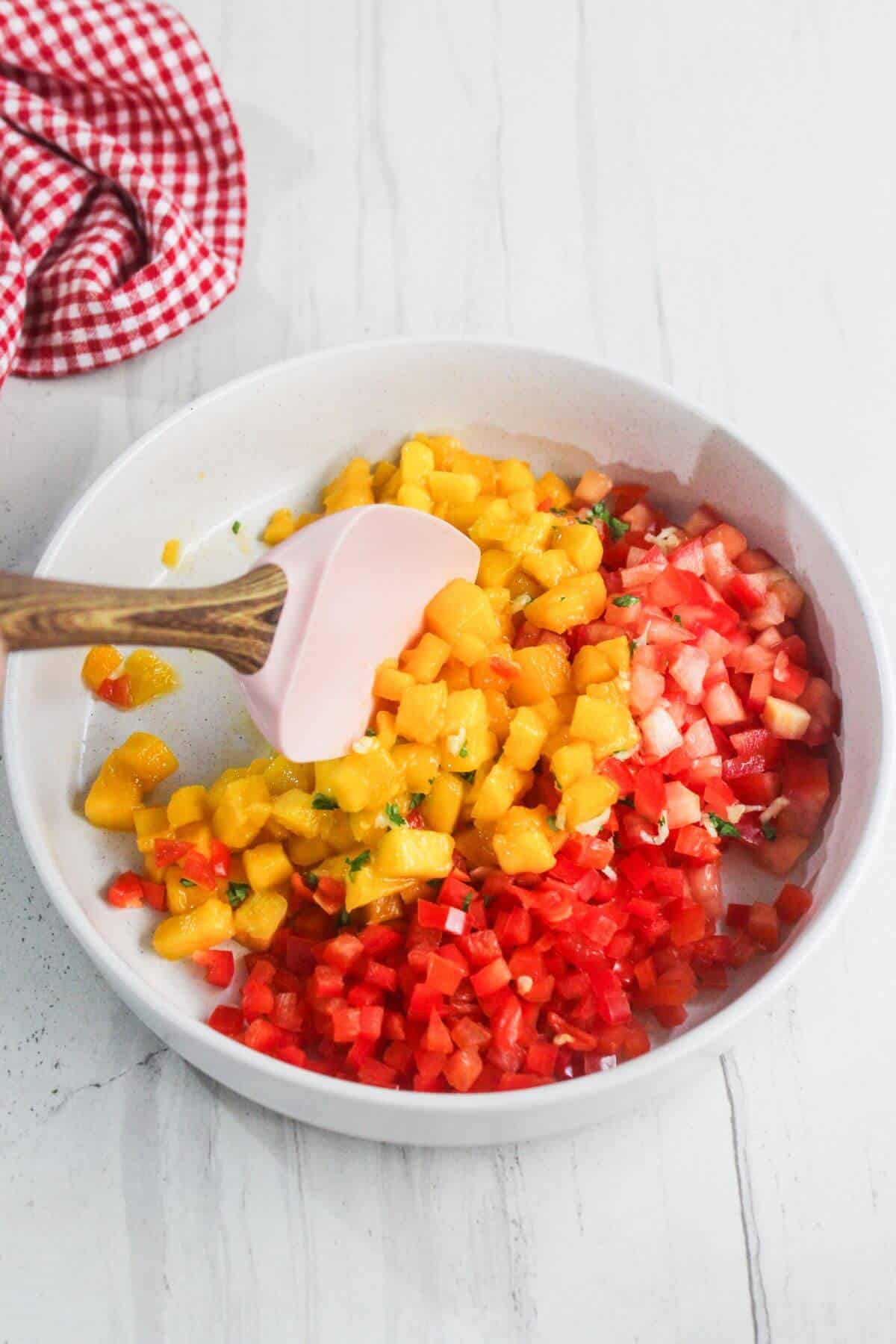 A white bowl contains diced yellow mango and red tomato, partially mixed with a pink spatula. A red and white checkered cloth is nearby on a white surface.