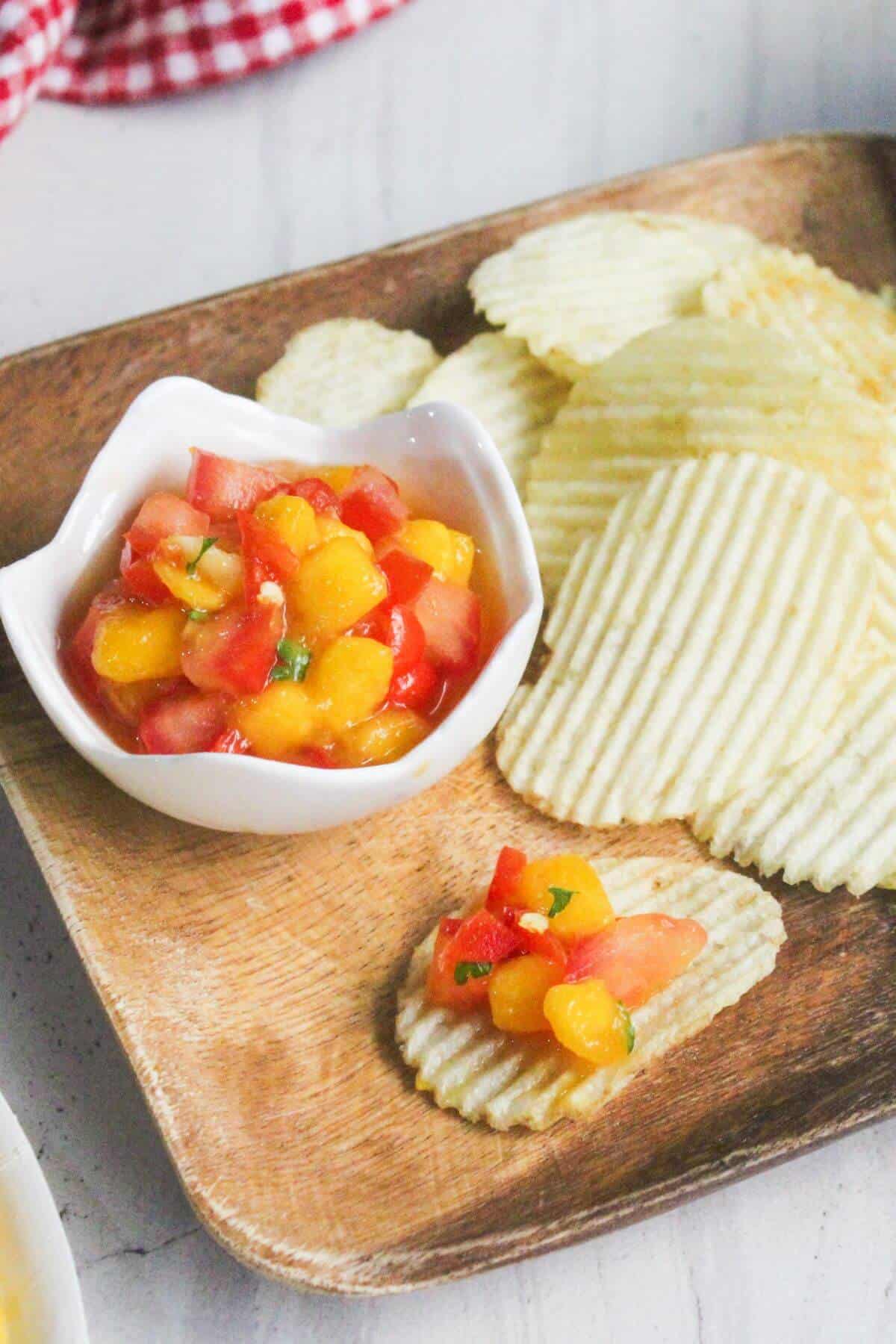 Ridged potato chips on a wooden tray with a small bowl of chunky peach and tomato salsa; one chip is topped with the salsa.