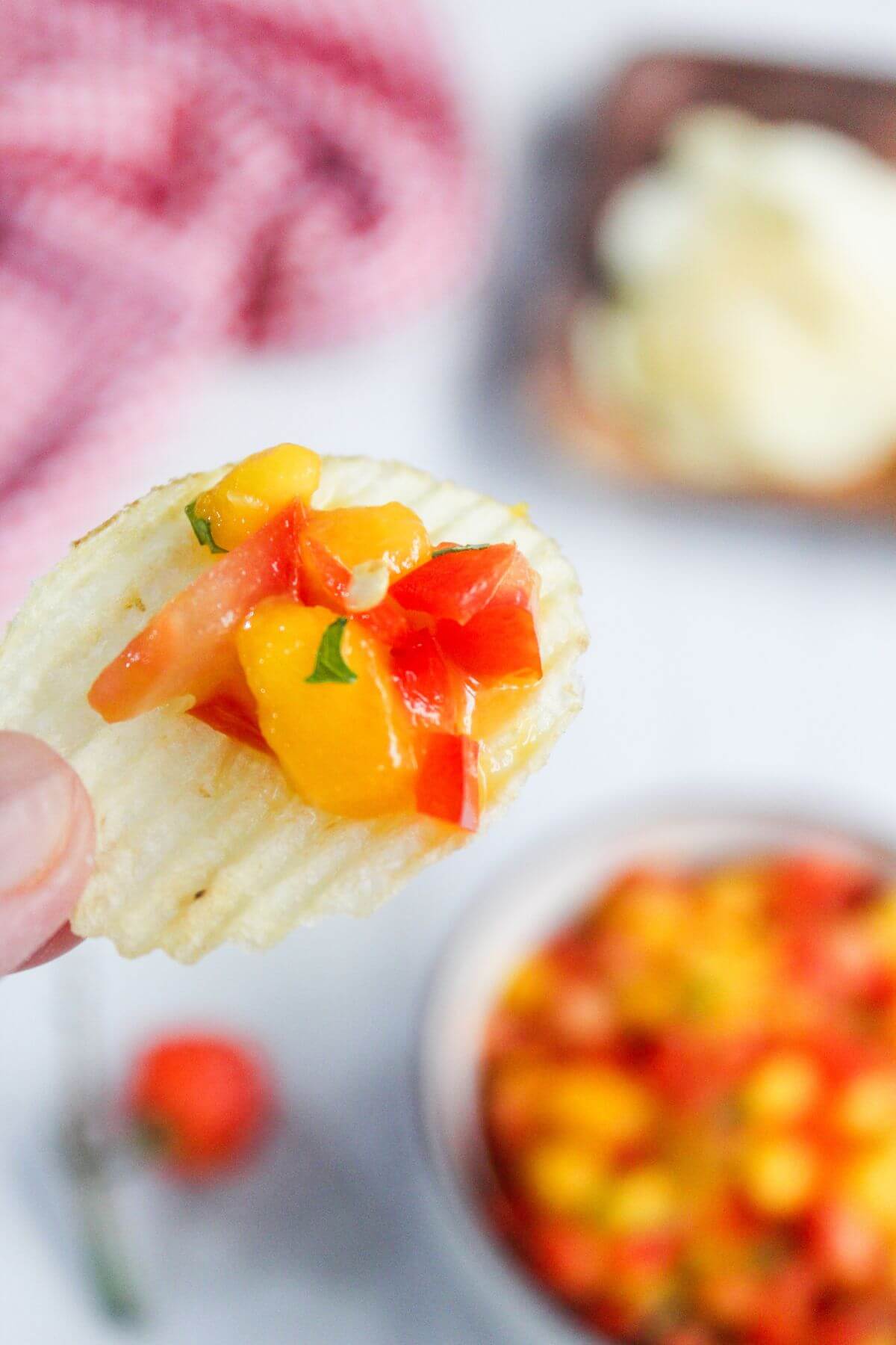 A ridged potato chip topped with diced mango salsa is held above a bowl of salsa, with a red and white cloth and another dish blurred in the background.