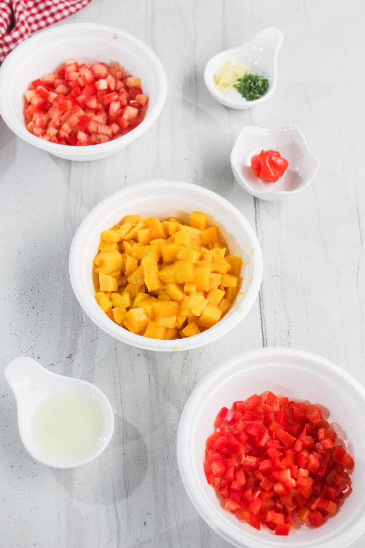 Bowls containing diced tomatoes, mango, red bell pepper, chopped herbs, minced garlic, a red chili pepper, and a small pitcher of lime juice on a white countertop.