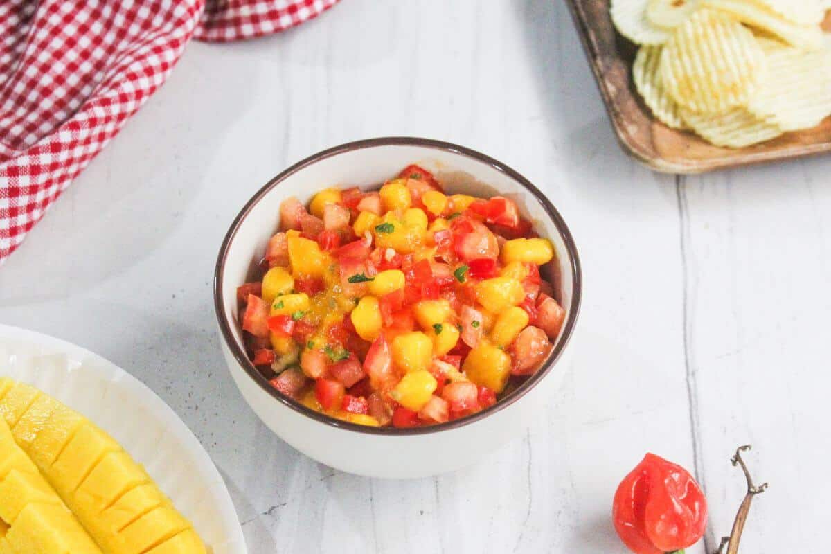 A bowl of mango salsa with diced tomatoes and herbs sits on a white surface, surrounded by sliced mango, potato chips, and a red checkered cloth.