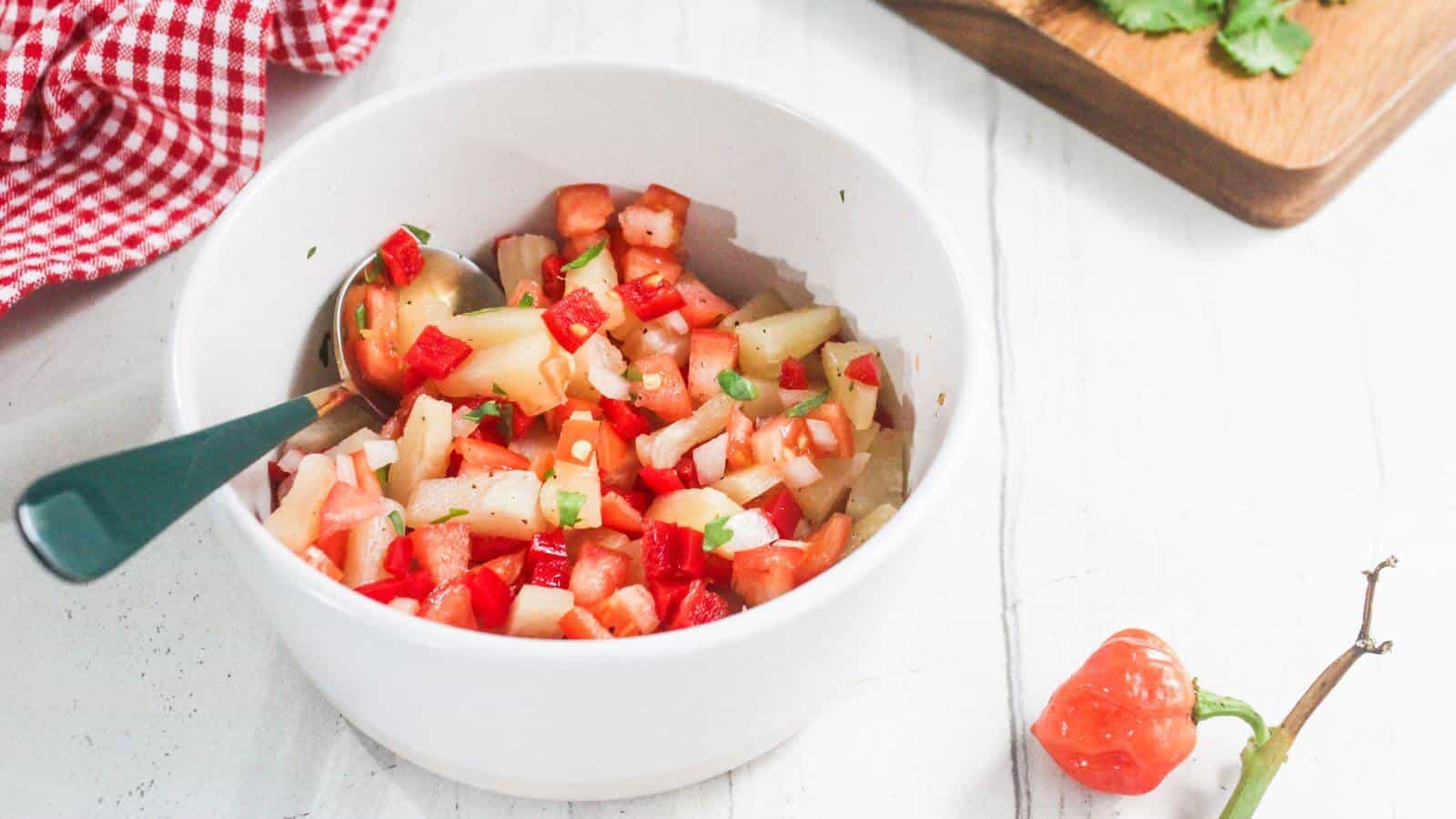A white bowl filled with diced tomatoes, onions, and peppers sits on a white surface, next to a chili pepper and a cutting board with cilantro.