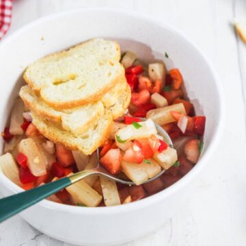 A white bowl with a vegetable salad containing diced tomatoes, onions, and red peppers, topped with three slices of bread. A spoon rests inside the bowl.