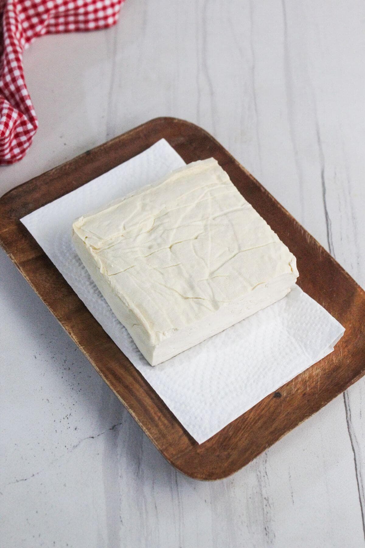 A block of tofu rests on a paper towel atop a wooden tray, with a red and white cloth in the background on a light-colored surface.