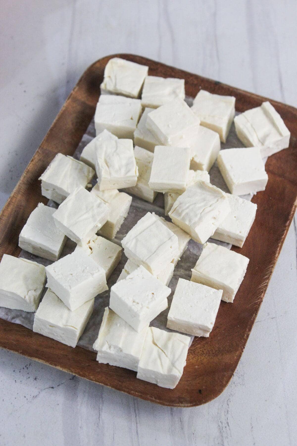 A wooden tray holds several rows of square, white marshmallows arranged on a piece of parchment paper.