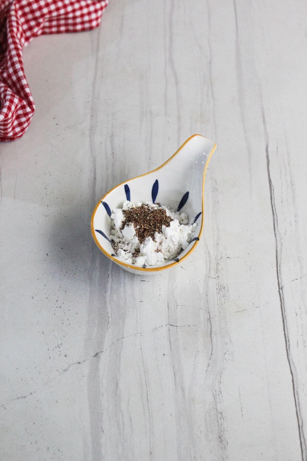 A small ceramic dish with blue patterns contains a mixture of white cheese and ground black pepper, set on a light gray marble surface beside a red checkered cloth.