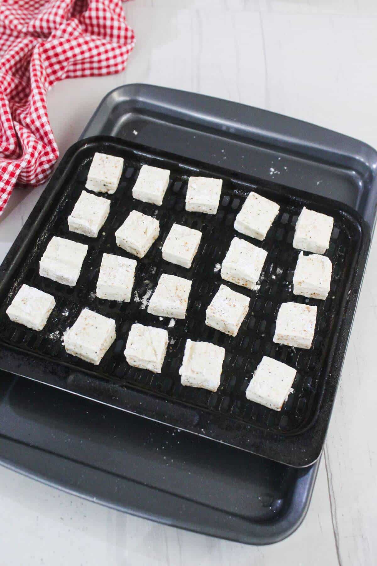 A black baking tray holds evenly spaced, uncooked tofu cubes dusted with seasoning. A red checkered cloth is partially visible in the upper left corner.