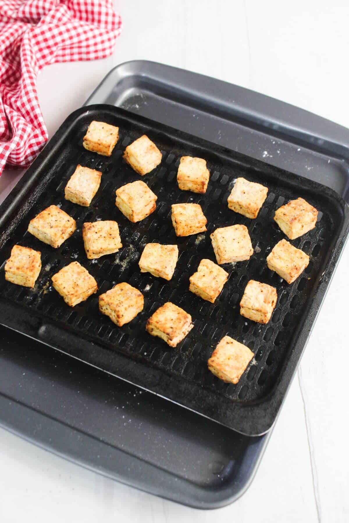 A baking tray with evenly spaced, golden-brown tofu cubes on a black grill rack, with a red checkered cloth in the background.
