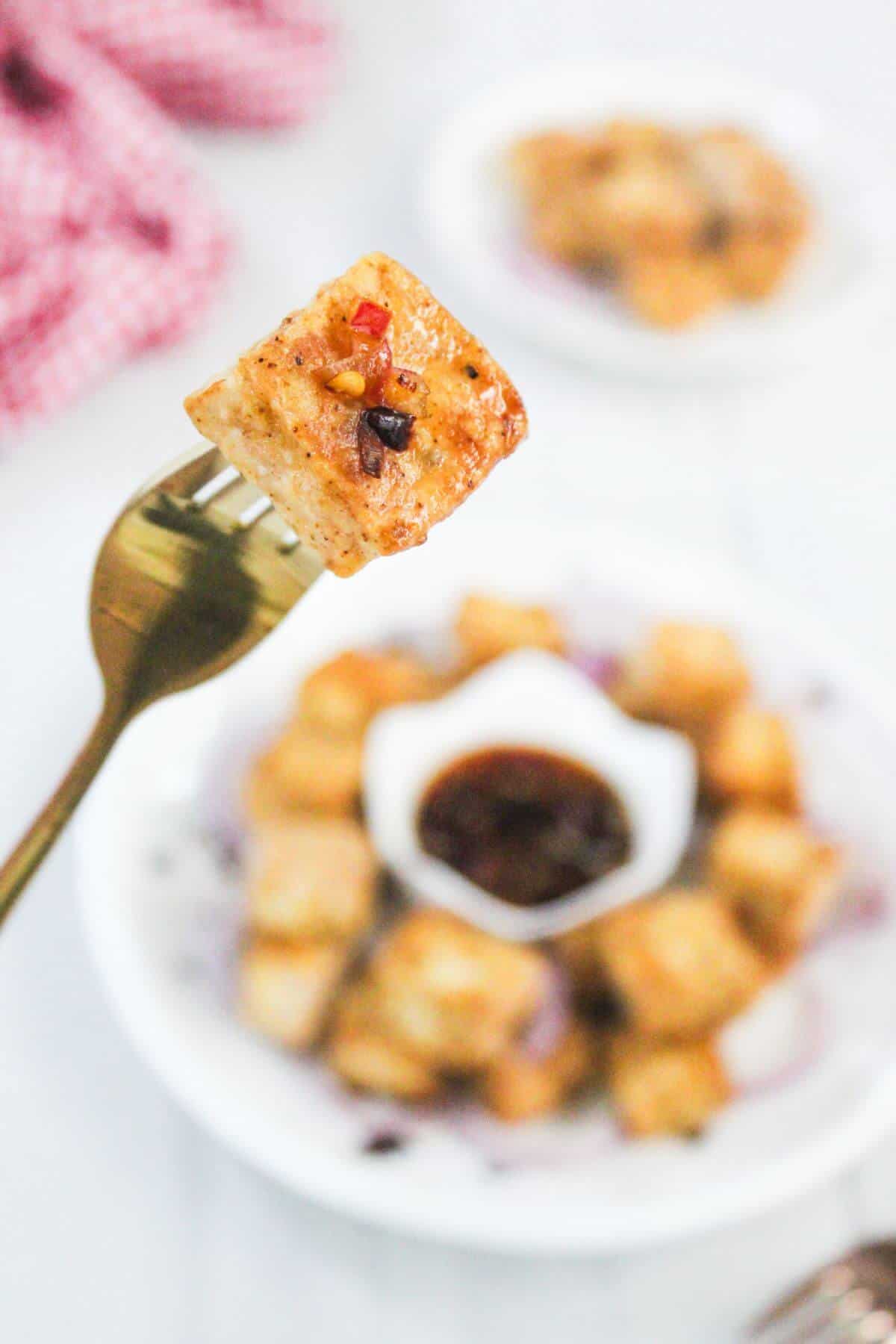 A fork holding a seasoned tofu cube above a plate of tofu cubes and a small bowl of dipping sauce.