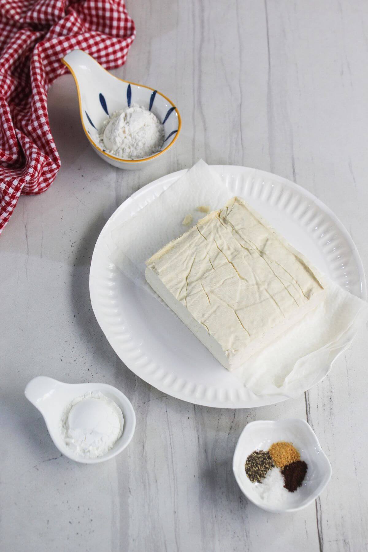Block of tofu on a white plate with parchment paper, surrounded by small dishes of spices and flour on a light surface with a red checkered cloth.