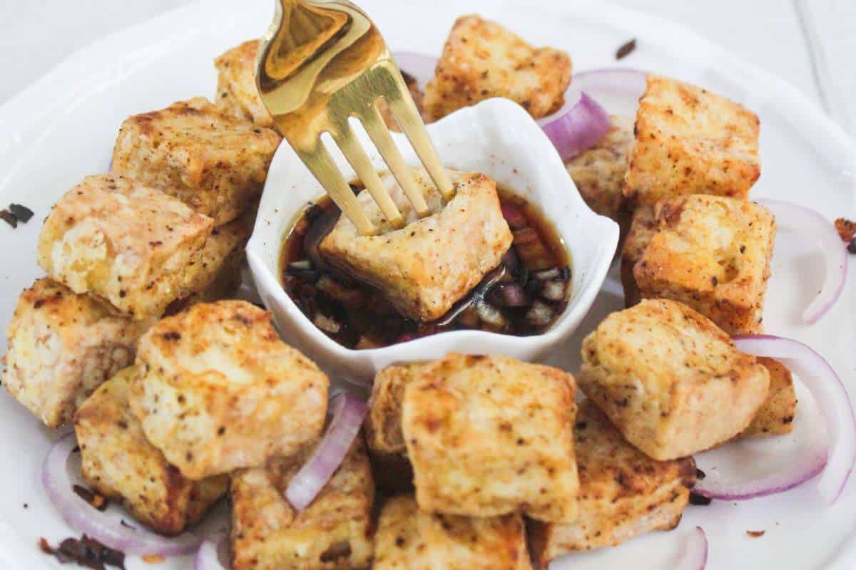 A plate of seasoned, baked tofu cubes arranged around a small bowl of soy sauce, with a fork picking up one piece and slices of red onion on the side.