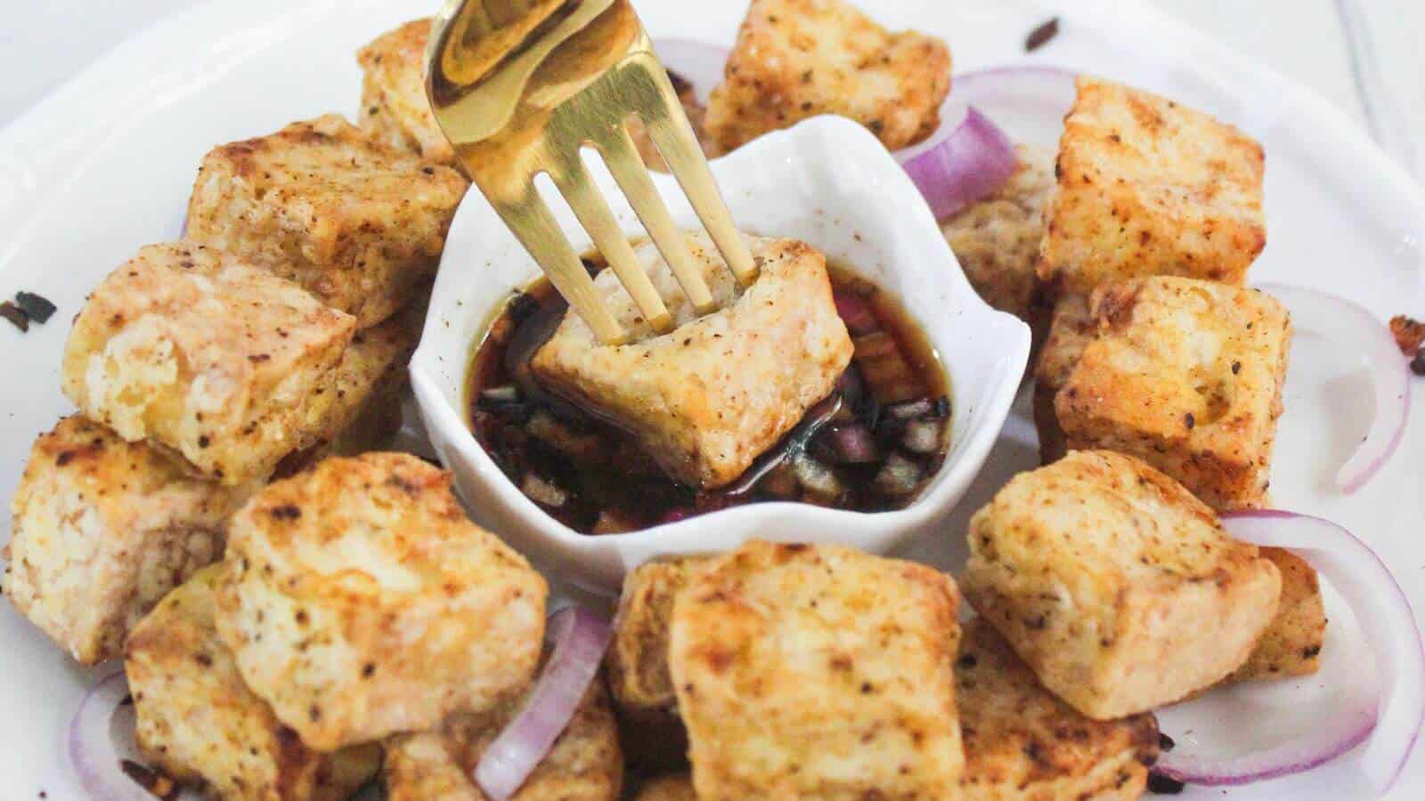 A plate of seasoned tofu cubes arranged around a small dish of soy sauce, with a fork dipping a tofu piece and slices of red onion on the plate.