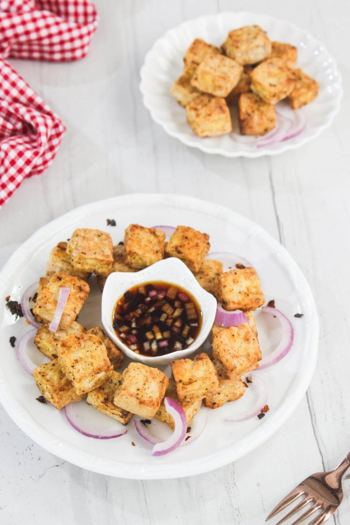 A plate of seasoned tofu cubes arranged around a bowl of dipping sauce, garnished with sliced red onions; extra tofu cubes are on a separate plate in the background.