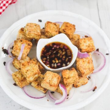 A plate of fried tofu cubes arranged in a circle with sliced red onions, surrounding a bowl of dipping sauce on a white plate.