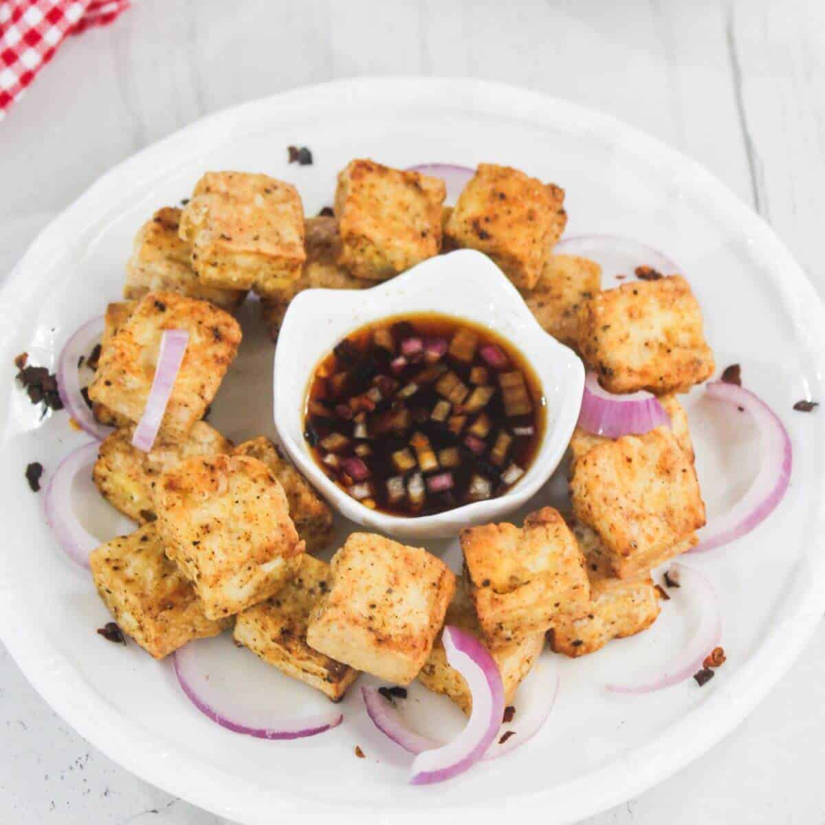 A plate of fried tofu cubes arranged in a circle with sliced red onions, surrounding a bowl of dipping sauce on a white plate.