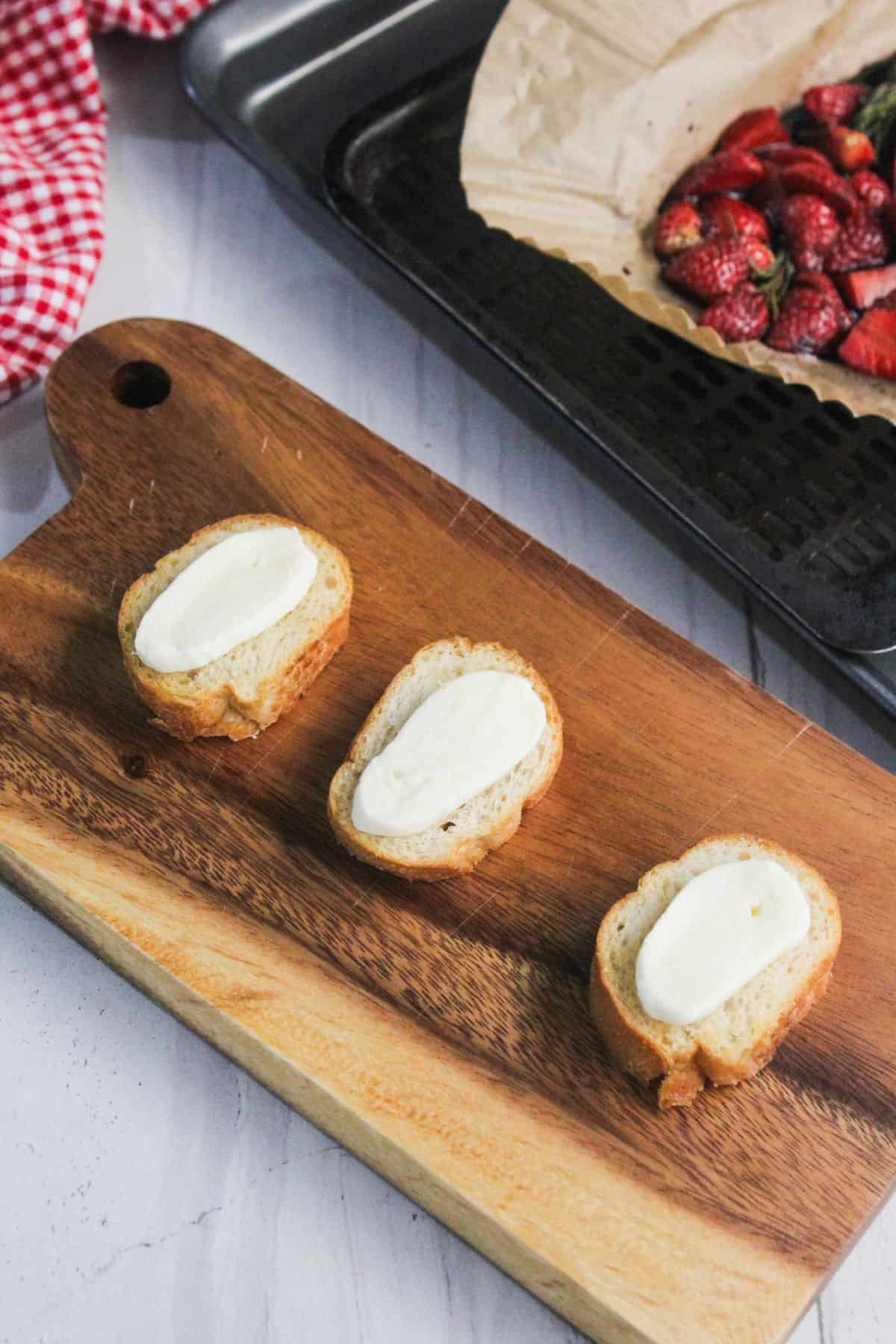 Three slices of bread topped with cream cheese are arranged on a wooden cutting board, with roasted strawberries in the background-perfect for creating delicious strawberry balsamic crostini.