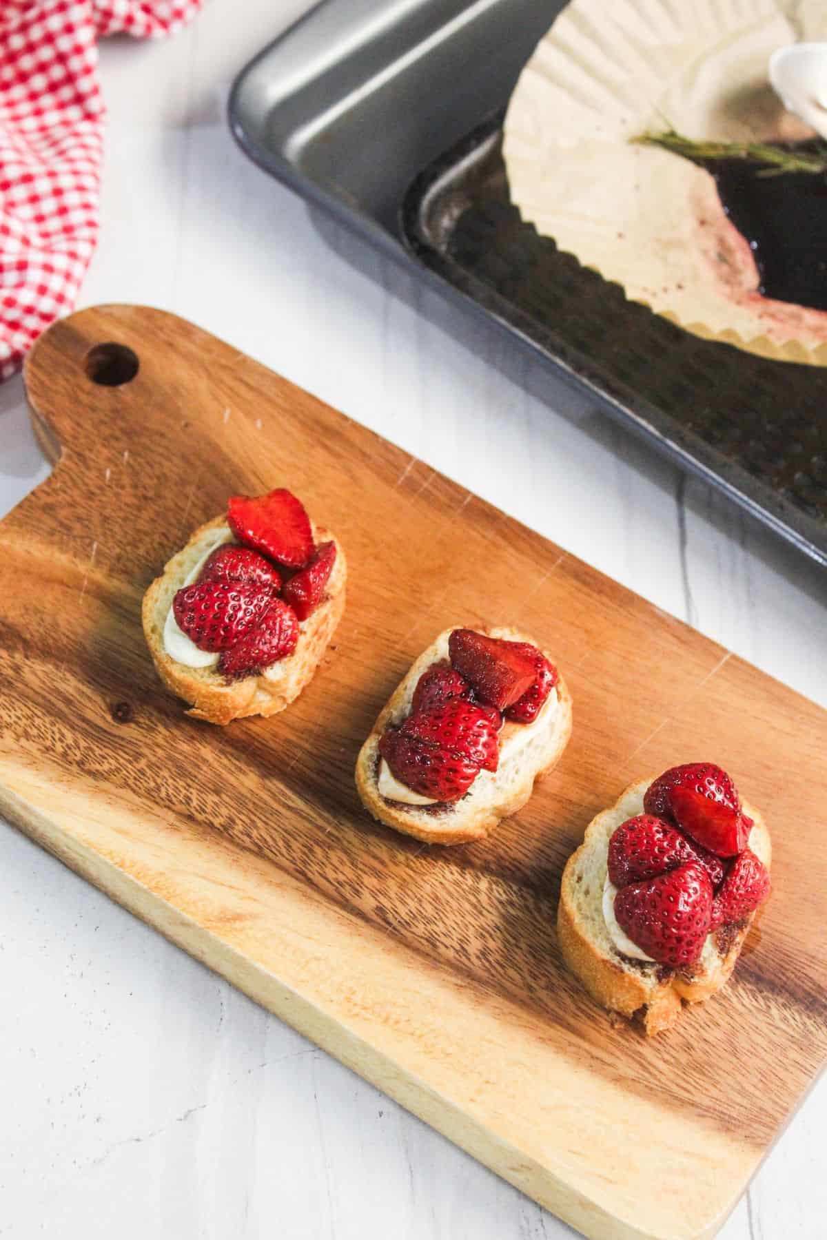 Three pieces of toasted bread, arranged on a wooden cutting board, are topped with cream and sliced strawberries for a delightful strawberry balsamic crostini.