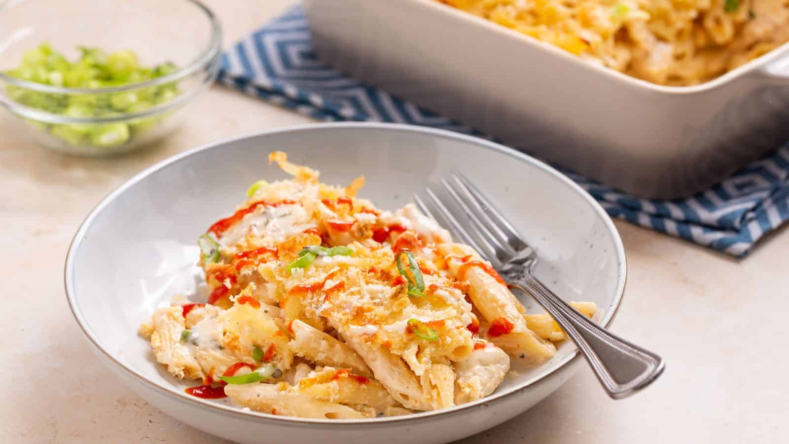 A bowl of baked pasta topped with sauce and chopped green onions, with a fork on the side. A baking dish and a bowl of green onions are in the background.