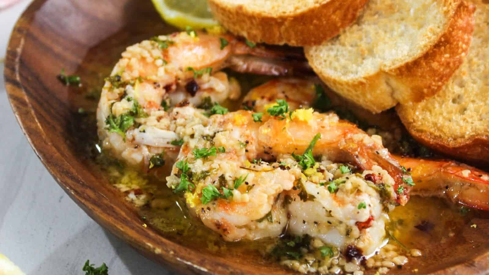 Cooked shrimp in a garlic butter sauce, garnished with chopped parsley, served in a wooden bowl alongside slices of toasted bread and a lemon wedge.