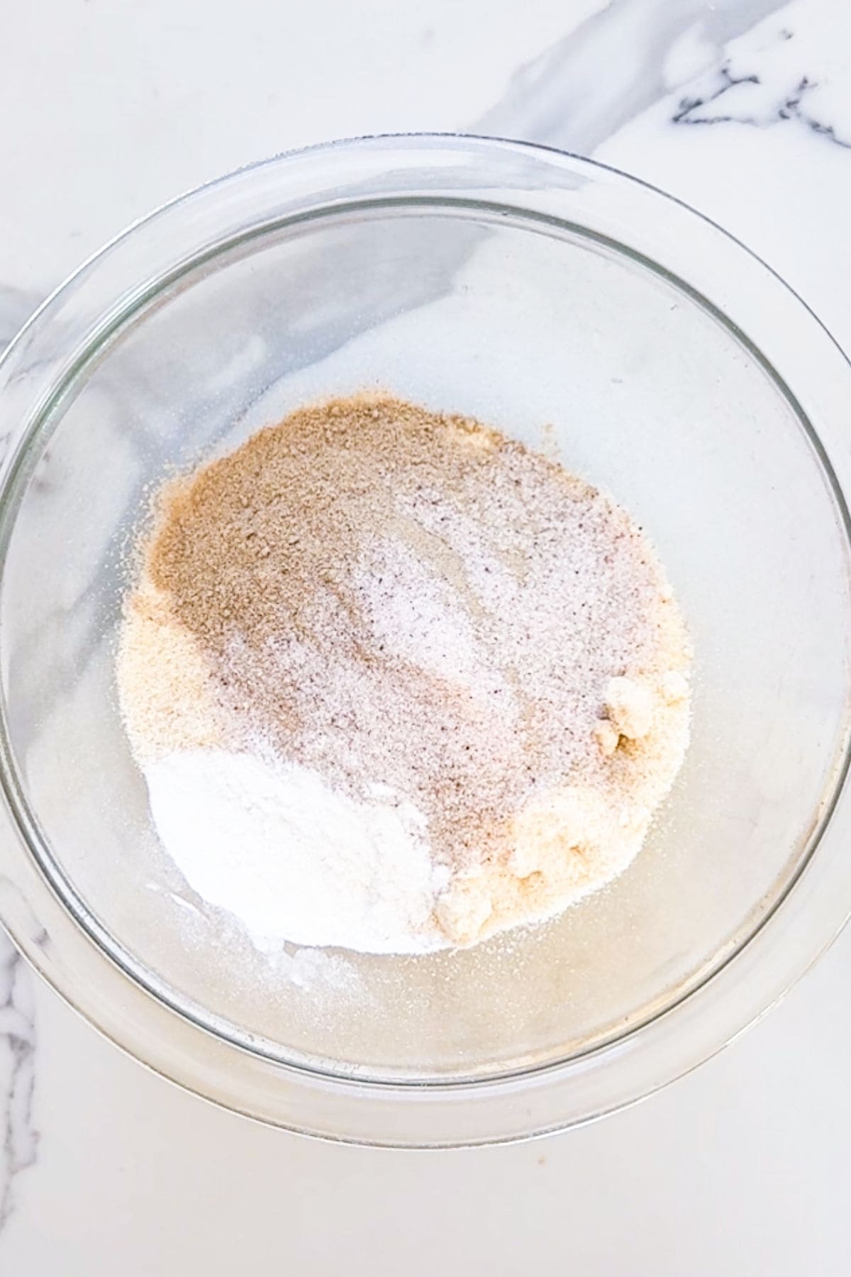 A glass bowl containing several types of dry ingredients, including white and brown powders, sits on a white marble surface.