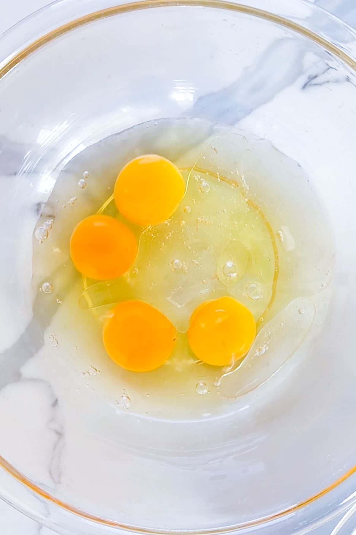 Four raw eggs with intact yolks in a clear glass bowl on a white marble surface.