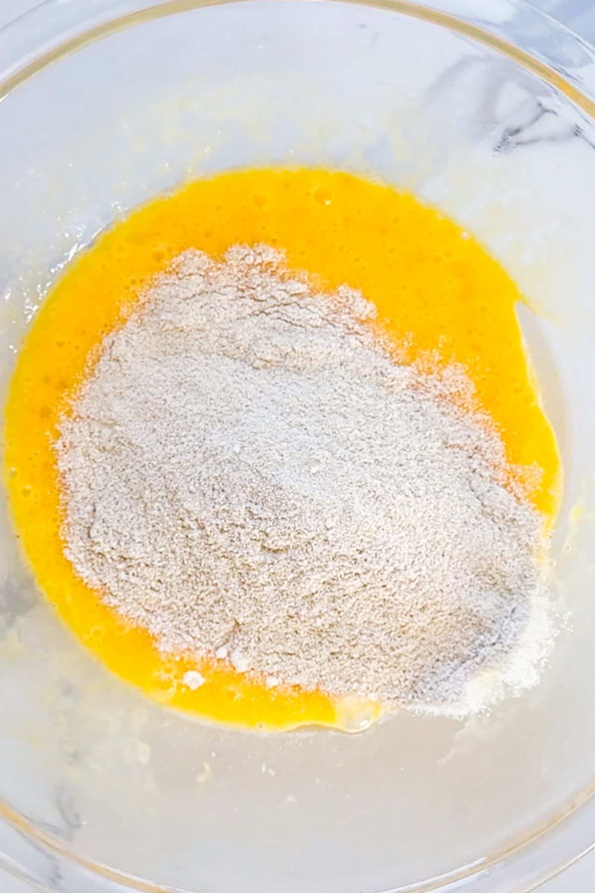 A glass bowl containing beaten eggs with flour or dry ingredients piled on top, ready to be mixed.