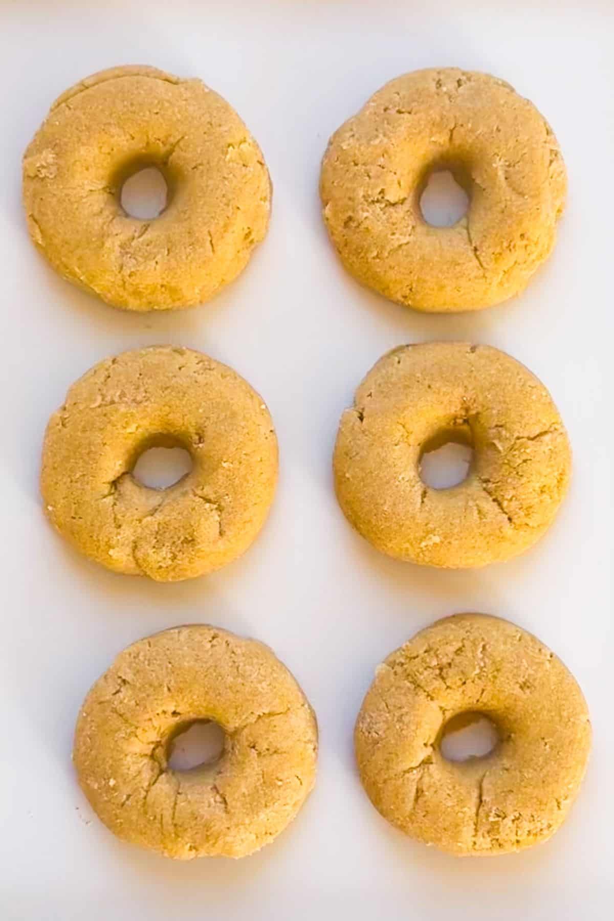 Six round, tan baked donuts with a cracked surface are arranged in two rows of three on a white background.