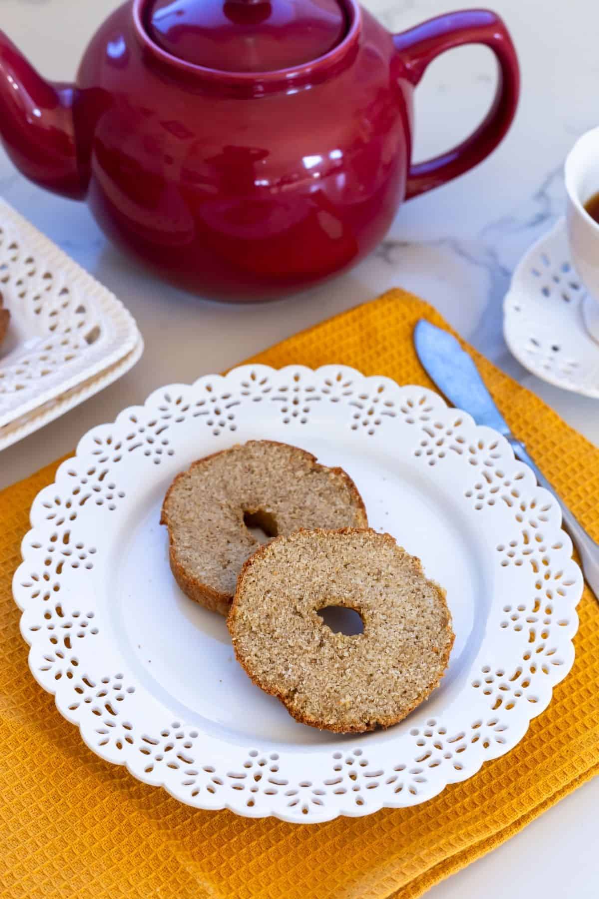 Two slices of a plain bagel on a white decorative plate, next to a red teapot and a cup, on a yellow placemat.