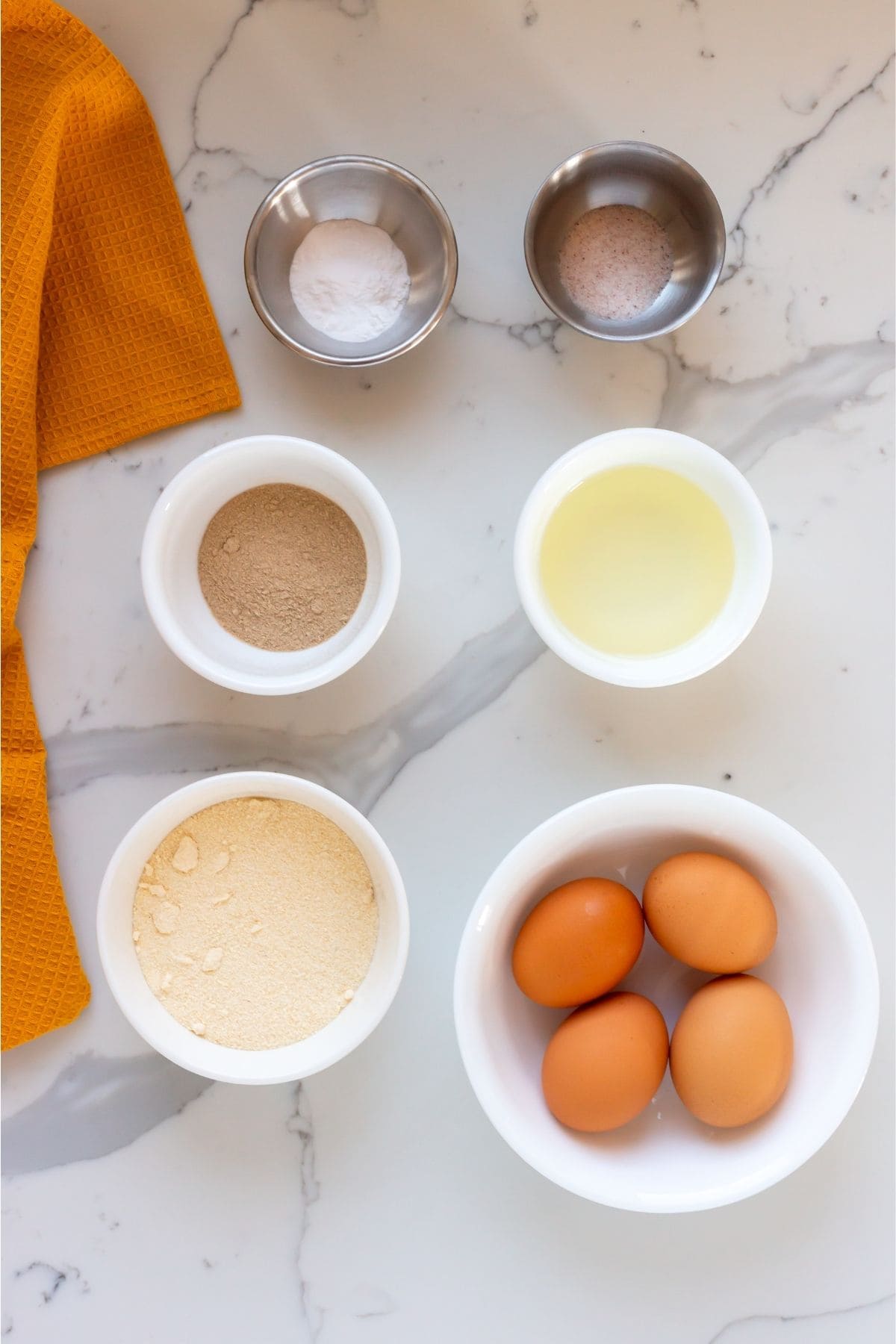 Bowls containing baking powder, salt, yeast, oil, flour, and four eggs are arranged on a marble countertop next to an orange kitchen towel.