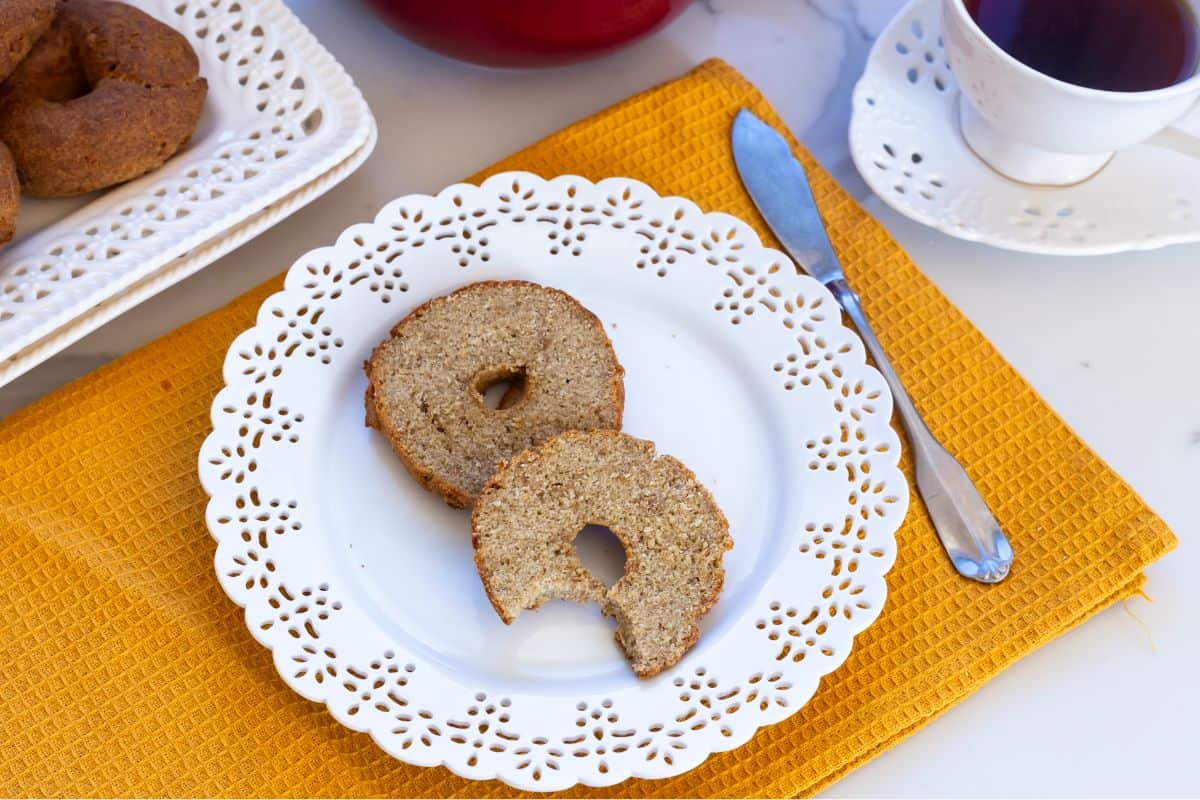Two bagel halves on a decorative white plate, one with a bite taken out, placed on a yellow mat next to a knife and a cup of tea.
