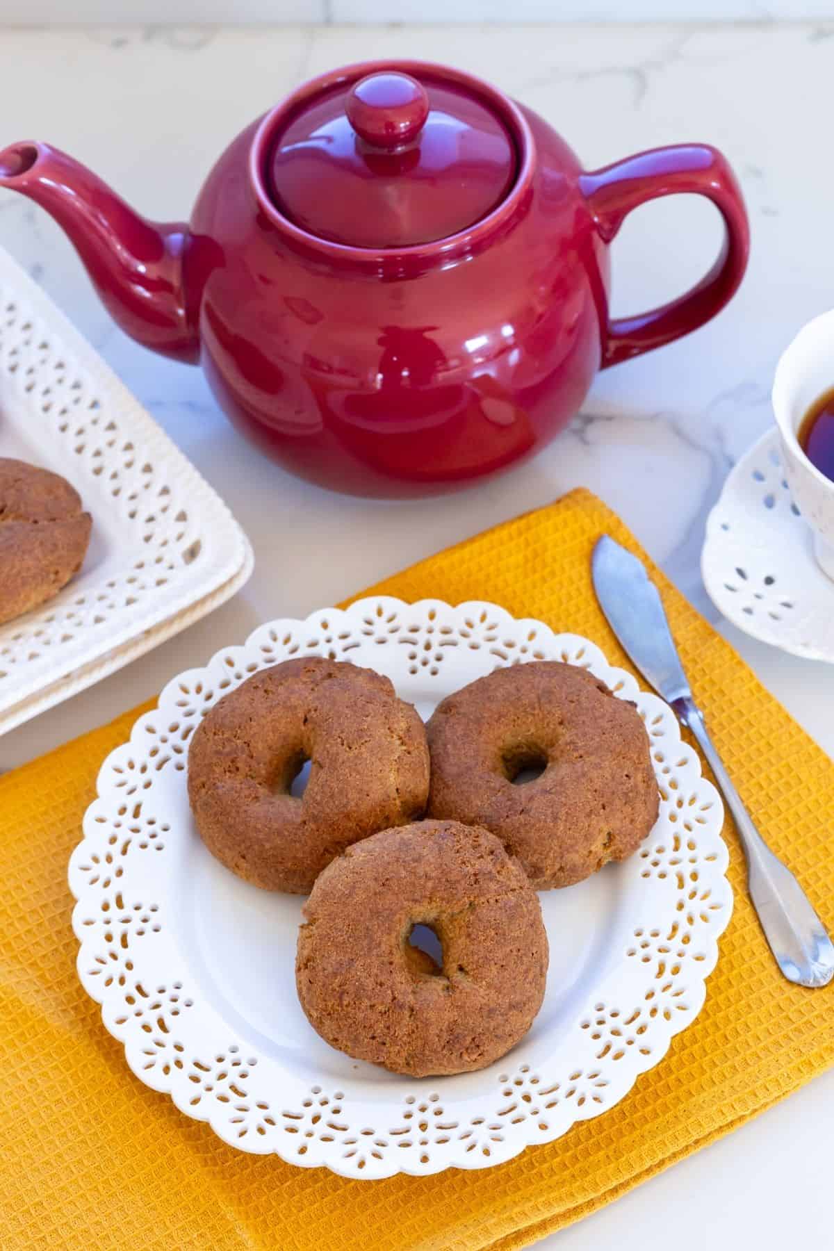 Three round donuts are on a white decorative plate with a yellow placemat, next to a red teapot, a butter knife, and a white cup on a marble surface.