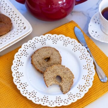Two bagel halves, one with a bite taken out, on a decorative white plate set on a yellow placemat, next to a knife, teacup, and red teapot.