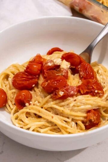 A white bowl of spaghetti with creamy sauce, roasted cherry tomatoes, and a fork, placed on a white countertop next to a white napkin.