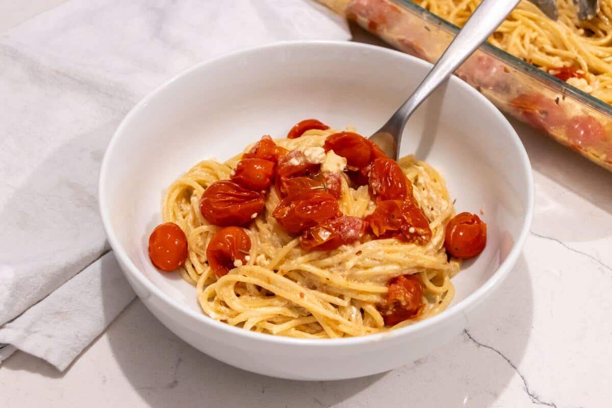 A white bowl filled with spaghetti, roasted cherry tomatoes, and a creamy sauce, with a fork resting in the bowl. A baking dish with more pasta is in the background.
