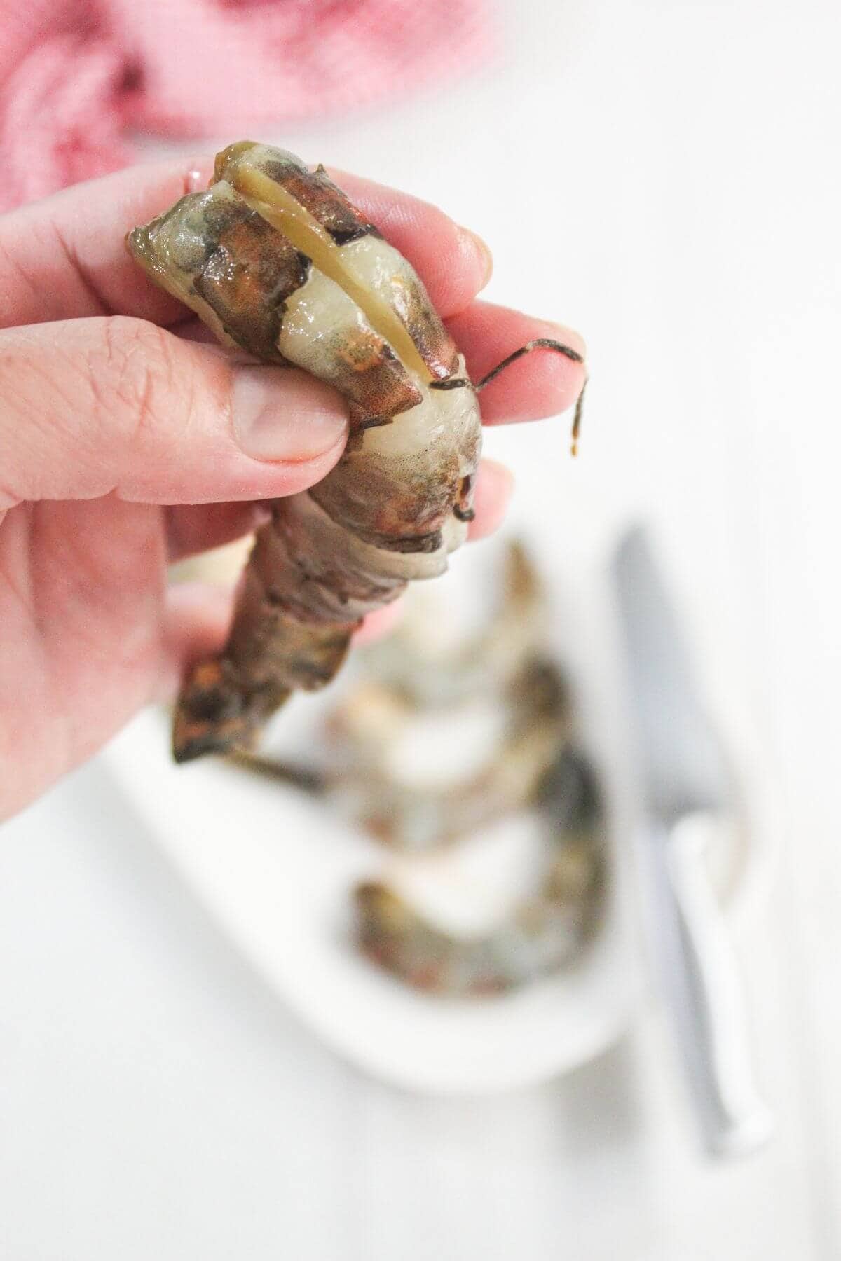 A hand holding a raw shrimp with its shell partially removed; two more shrimp and a knife are on a plate in the background.