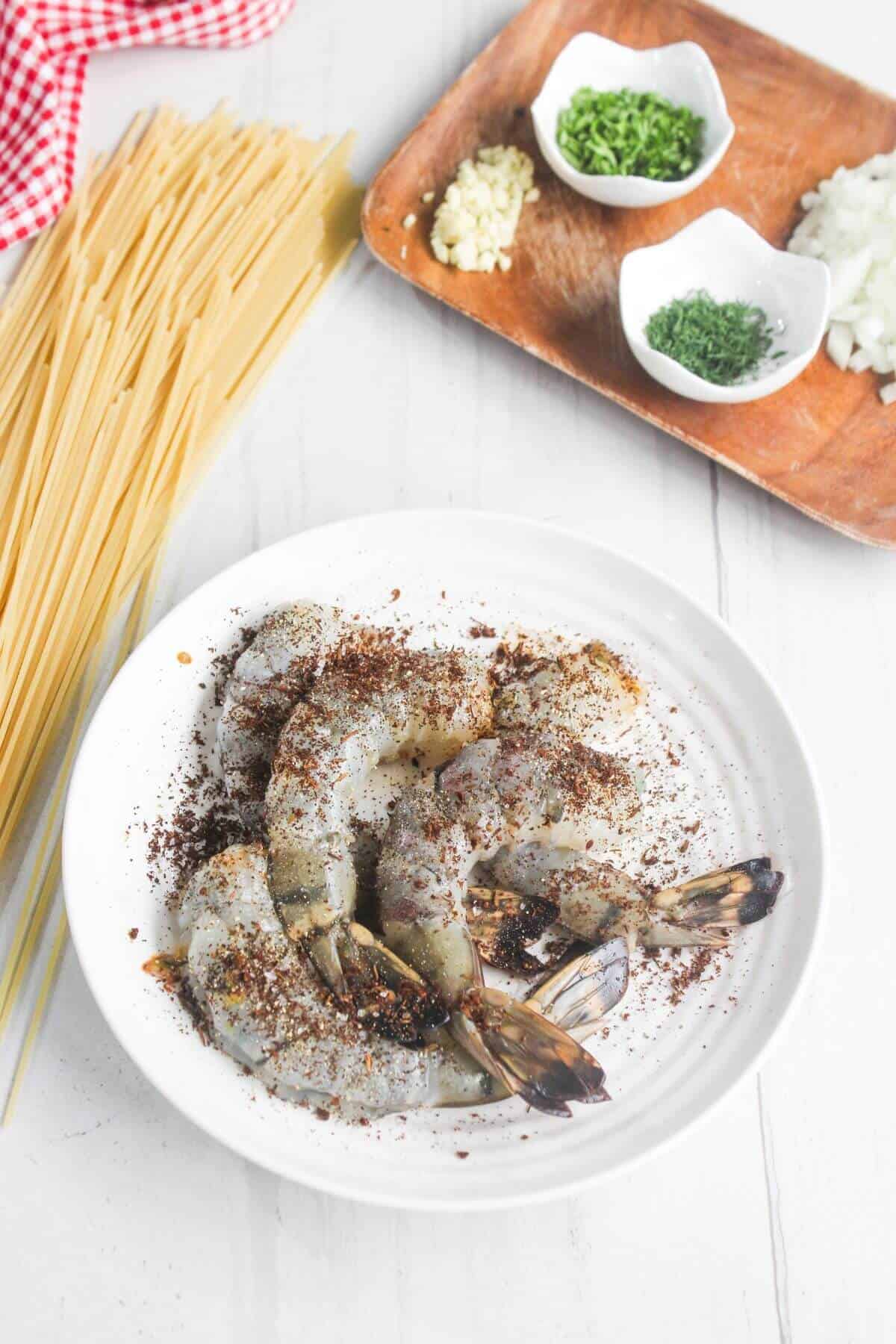 Raw shrimp seasoned with spices on a white plate, uncooked spaghetti beside it, and chopped herbs, garlic, and onions on a wooden tray in the background.