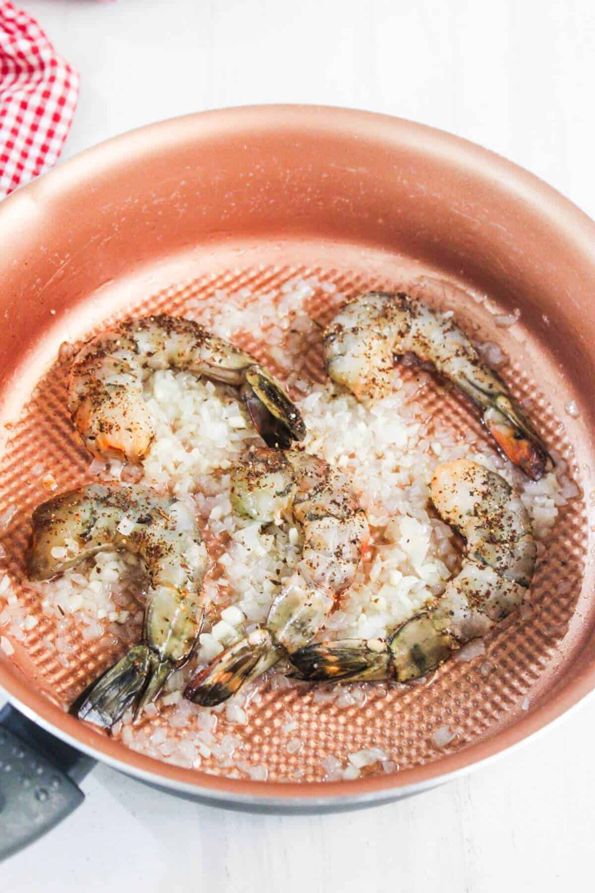 Raw shrimp and chopped onions are being cooked in a copper frying pan, seasoned with black pepper.
