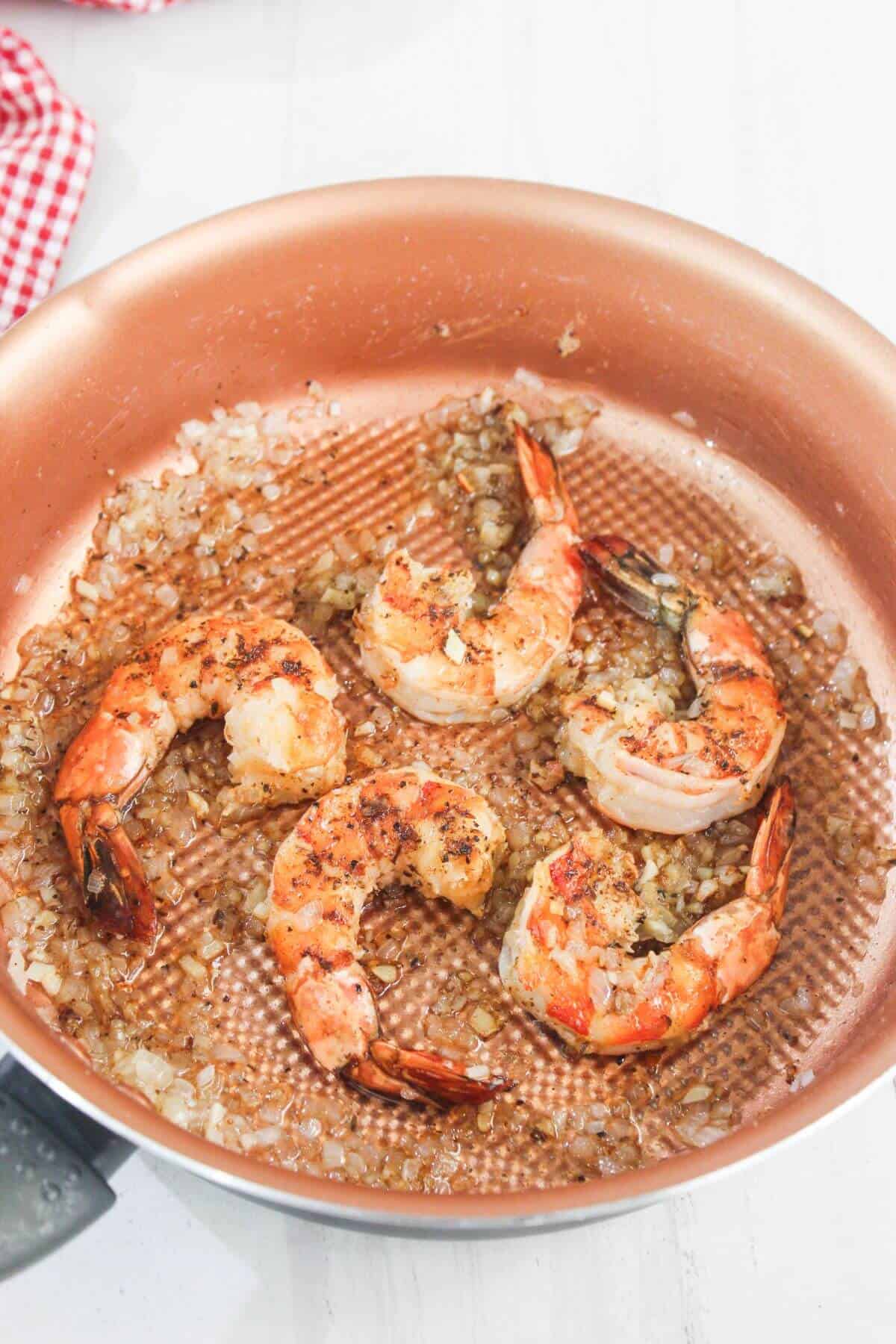 Four cooked shrimp with diced onions in a copper frying pan on a white surface.
