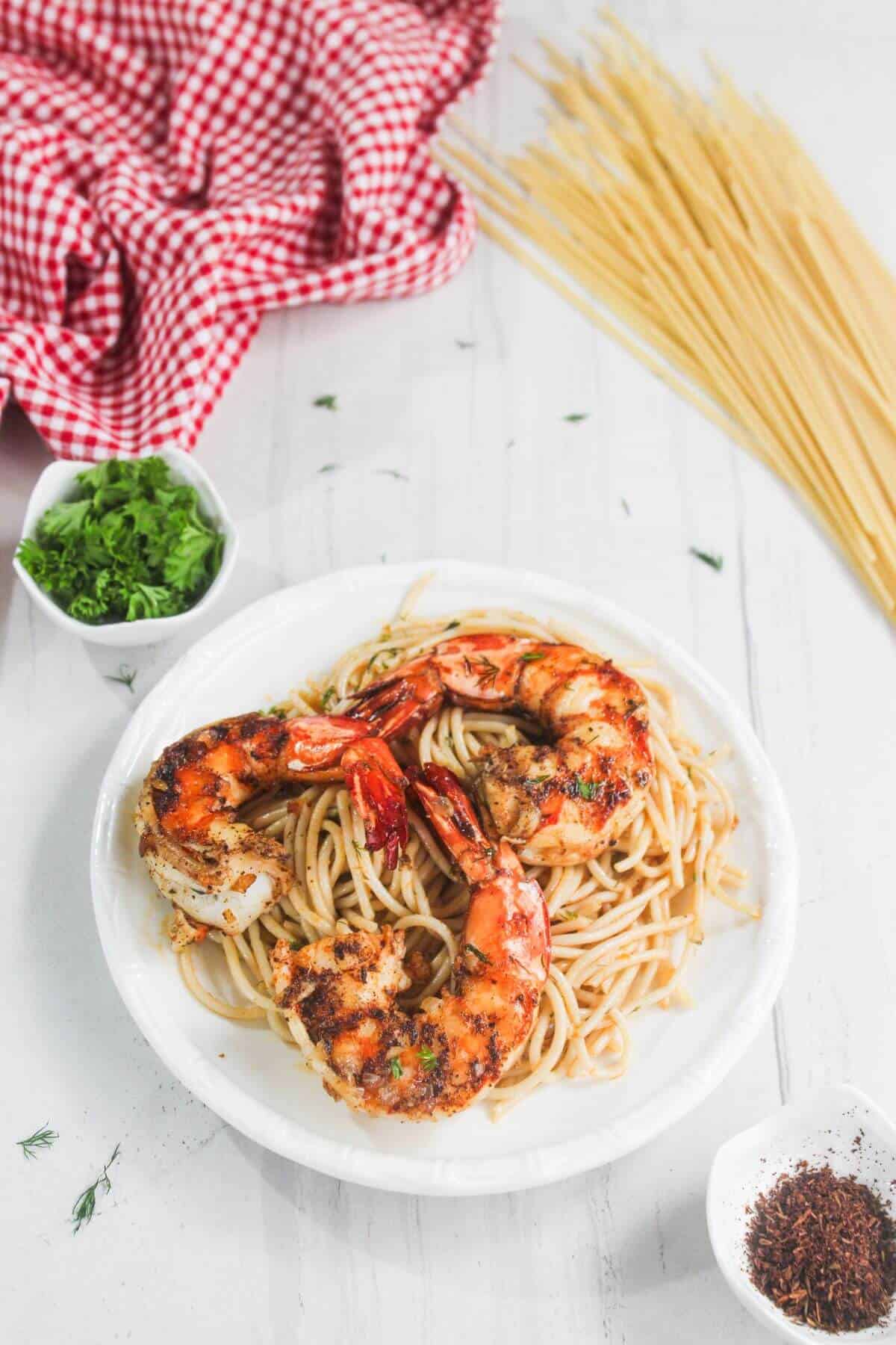 A plate of spaghetti topped with grilled shrimp, surrounded by uncooked spaghetti, chopped herbs in a bowl, chili flakes, and a red checkered cloth on a white table.