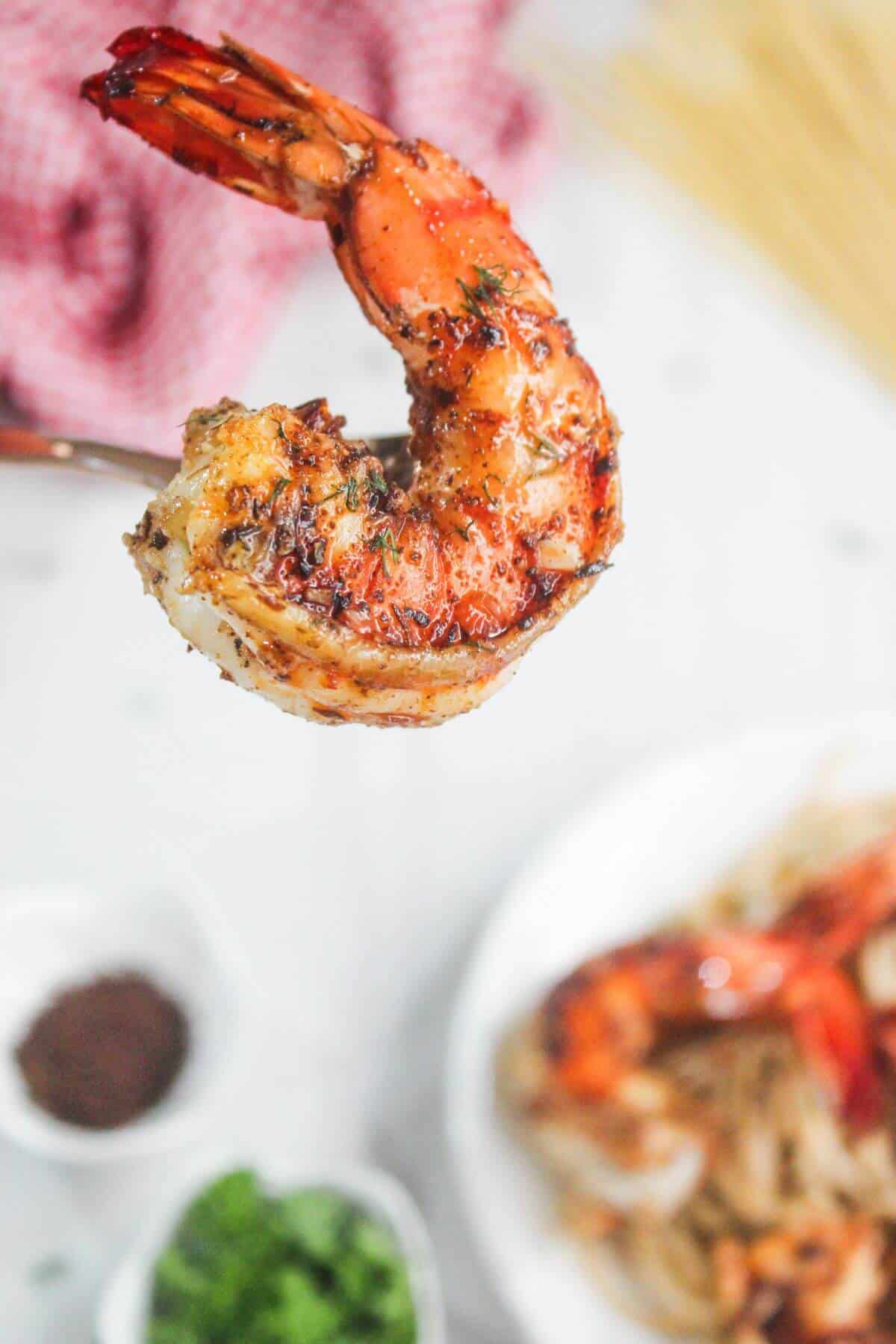 Close-up of a grilled shrimp held by a fork, with a plate of pasta and another shrimp blurred in the background.