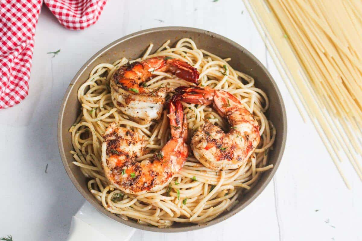 A skillet filled with cooked spaghetti topped with three large, seasoned shrimp. Uncooked spaghetti and a red checkered cloth are nearby on a white surface.